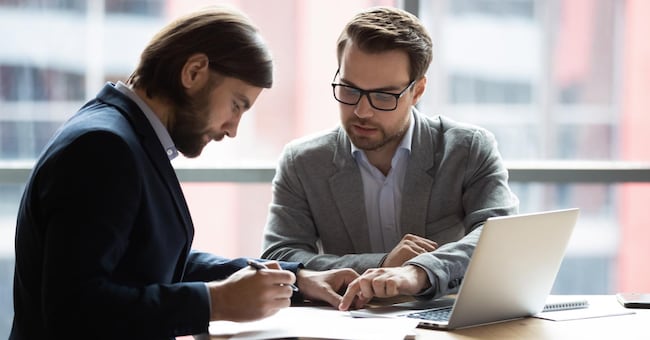 Focused young businessman signing agreement with skilled lawyer in eyeglasses. Concentrated financial advisor showing place for signature on paper contract document to male client at meeting in office