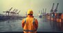 A port worker from behind with a safety vest and protective helmet looks into the harbor basin with containers.