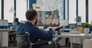 Back view of handicapped paralysed invalid manager talking with team on videocall about sale report, having video conference working in start-up office. Team using online meeting discussing on webcam