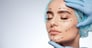 Beauty portrait of woman with closed eyes, plastic surgery concept, studio. Head and shoulders of model in blue cap with puncture lines on face, hands in blue gloves holding face