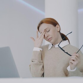 Employee has headache, touching her eyes. Forehead pain. Tired young woman at the desk in office. Girl is holding her head with hands. Concept of overworking and pressure at workplace.