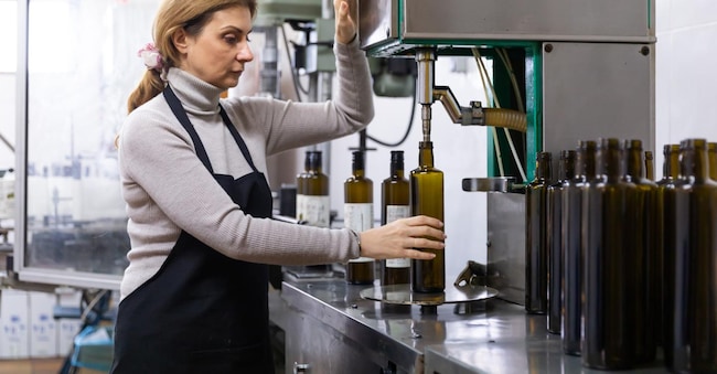 Skilled woman engaged in olive oil production, controlling process of bottling of finished product in glass bottles