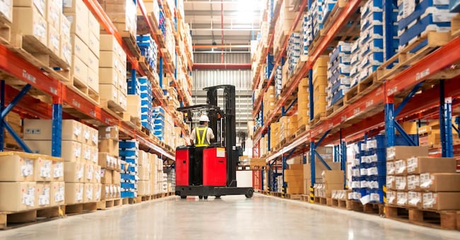 Worker in forklift-truck loading packed goods in huge distribution warehouse with high shelves.
