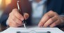 The photo is a close-up of a person's hand signing a document. The pen is in focus and the rest of the image is blurred. The person is wearing a suit jacket and a white shirt. The photo is taken from a low angle. The photo conveys a sense of professionalism and focus.