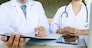Two doctors discussing patient notes in an office pointing to a clipboard with paperwork as they make a diagnosis or decide on treatment.