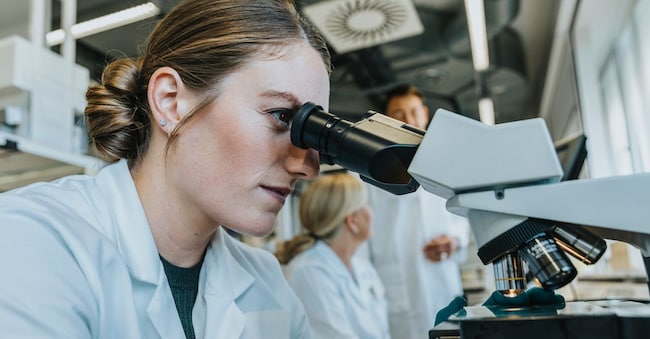 Assistant analyzing human brain microscope slide while sitting with scientist in background at laboratory
