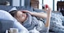 Sick exhausted girl in scarf is lying in bed wrapped in blanket. Young woman with fever and headache is measuring temperature with thermometer, treated at home. Winter cold and flu concept.