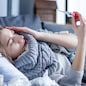 Sick exhausted girl in scarf is lying in bed wrapped in blanket. Young woman with fever and headache is measuring temperature with thermometer, treated at home. Winter cold and flu concept.