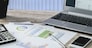 Modern workplace at the office: printouts of charts and financial data next to a laptop, smartphone and calculator on the desk.