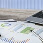 Modern workplace at the office: printouts of charts and financial data next to a laptop, smartphone and calculator on the desk.