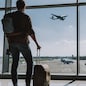 Guy is standing with luggage near window in waiting room. He is glancing at plane taking off outside. Copy space in right side