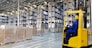 Huge distribution warehouse with high empty shelves and forklift with driver in yellow helmet.