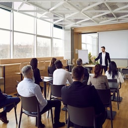 Team of people having class with business trainer. Group of male and female employees sitting at desks in modern office and listening to lecture by experienced teacher sharing knowledge and guidance