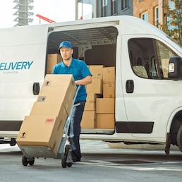 Delivery Man Pushes Hand Truck Trolley Full of Cardboard Boxes Hands Package to a Customer. Courier Delivers Parcel to Man in Business District.