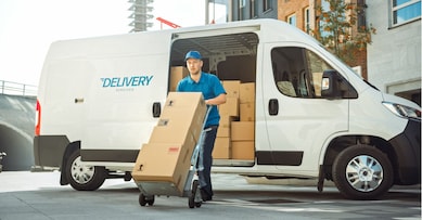 Delivery Man Pushes Hand Truck Trolley Full of Cardboard Boxes Hands Package to a Customer. Courier Delivers Parcel to Man in Business District.