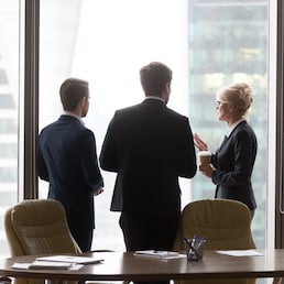 Multiracial workers colleagues talk at break, drink coffee, stand near window, modern city skyscrapers. Coworkers in office discuss future project, planning new opportunities. Business vision concept