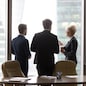 Multiracial workers colleagues talk at break, drink coffee, stand near window, modern city skyscrapers. Coworkers in office discuss future project, planning new opportunities. Business vision concept