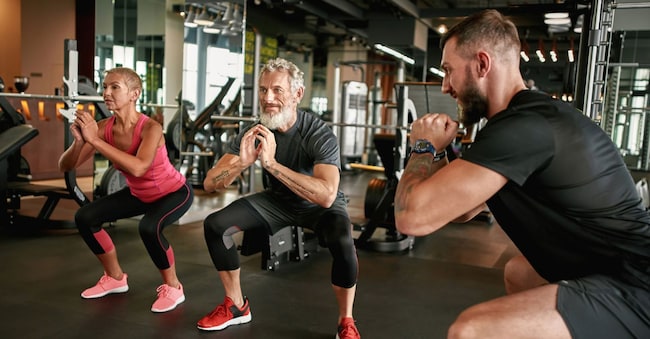 Attractive and fit elderly woman and man performing together injury free squats under professional trainer supervision. Excercises to strengthen body. Seniors exercising together concept. Svitlana - stock.adobe.com