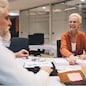 Senior woman in orange sweater smiles while discussing work with older male colleague in an office environment. Documents, charts, and notes spread across the table in a collaborative setting