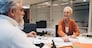 Senior woman in orange sweater smiles while discussing work with older male colleague in an office environment. Documents, charts, and notes spread across the table in a collaborative setting