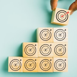 Close-up of a man's hand arranging wooden blocks in a stair-like formation, placing a cube with a target icon on top. Depicting growth, success, and strategic concepts. Isolated on a blue background.