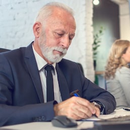 Senior business man wearing formalwear writing on paper in front of computer in office.