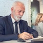 Senior business man wearing formalwear writing on paper in front of computer in office.