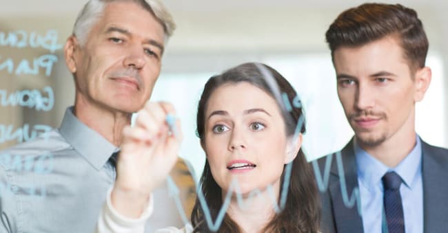 Serious young businesswoman drawing graph on transparent glass, young and senior male colleagues standing behind and looking at it with blurry office interior in background
