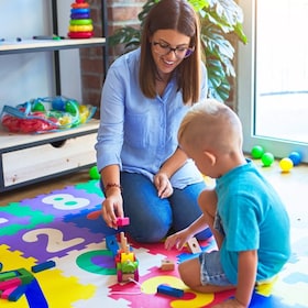 Young caucasian child playing at playschool with teacher. Mother and son playing with wooden pieces train at playroom