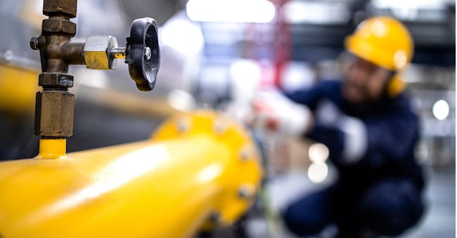 Close up view of natural gas pipeline with valve and refinery worker working in the background.