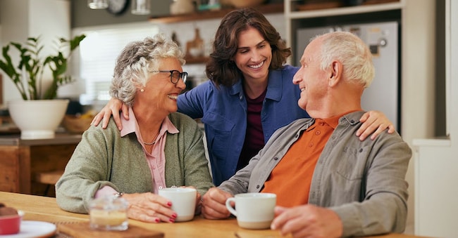 Senior father and old mother sharing tea and heartfelt conversation with daughter at home at tea time. Elderly parents and beautiful daughter bonding over tea and discussion. Warm daughter embracing from behind her joyful parents while sitting at table and having a good conversation. Rido - stock.adobe.com
