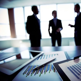 Close-shot of a tablet computer displaying financial data, three businessmen standing in the background