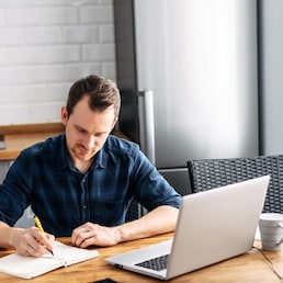 Online training. Young guy student studying at home sitting in the kitchen and using laptop. He writes with a pen in a notebook