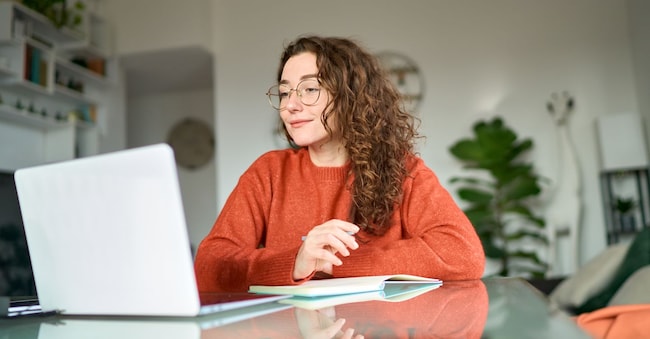Young female student using laptop studying online, remote working at home office using laptop computer watching webinar, elearning web training, having work video call sitting at table, writing notes.