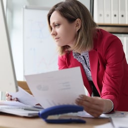 Focused woman compares paper accounting documents with computer version standing at table with big monitor. Female accountant does paperwork in light office