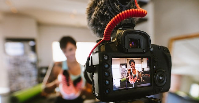 Female vlogger holding sports shoes in hands happily looking in camera while recording video for blog. Focus on camera with social media influencer reviewing and marketing a sports shoe.