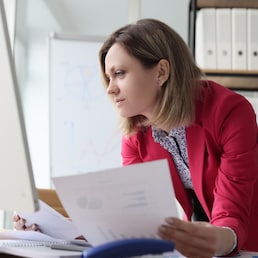 Focused woman compares paper accounting documents with computer version standing at table with big monitor. Female accountant does paperwork in light office