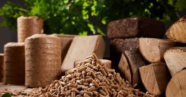 Stack of wood pellets arranged on table near chopped woodpile and compressed biomass briquettes against lush green tree in daytime