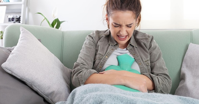 2SBFNPT Young woman sitting on sofa at home suffering from period pain, cramps. Holding hot water bottle on abdomen, dealing with endometriosis, indigestion, Alamy Stock Photo