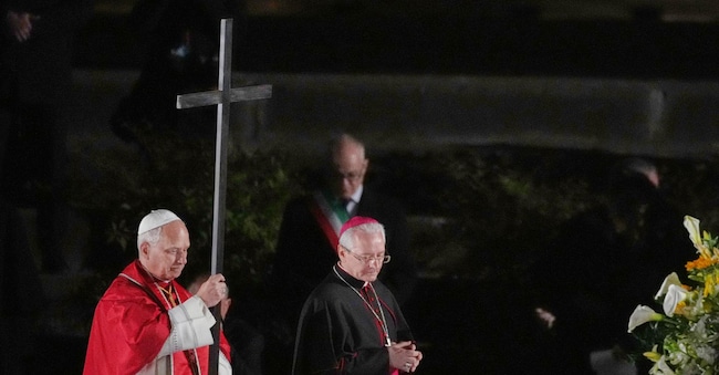 Pope Leo XIV, accompanied by Master of Pontifical Liturgical Celebrations, Archbishop Diego Giovanni Ravelli, right, carries a lightweight, 1.5-meter (5-foot) wooden cross during the Via Crucis, the torchlit Good Friday Stations of the Cross procession at the Colosseum in Rome, Friday, April 3, 2026, which symbolically retraces Jesus Christ's steps to his crucifixion on Calvary in Jerusalem. (AP Photo/Alessandra Tarantino) APN