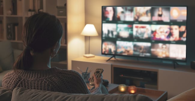 Una donna è distesa sul divano; sta usando il telecomando per scegliere un programma o un film dal menu della TV StockPhotoPro - stock.adobe.com