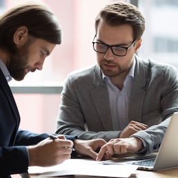 Focused young businessman signing agreement with skilled lawyer in eyeglasses. Concentrated financial advisor showing place for signature on paper contract document to male client at meeting in office fizkes - stock.adobe.com