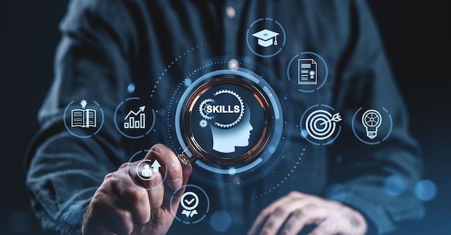 Person holding magnifying glass over digital icons symbolizing skills and learning, modern graphic style, dark blurred background, concept of professional development and education ImageFlow - stock.adobe.com