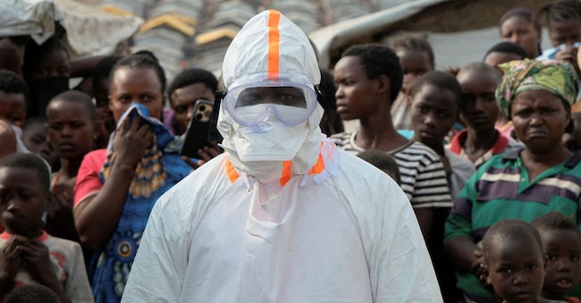 Un operatore sanitario in equipaggiamento di protezione individuale (DPI) si trova accanto a sfollati in attesa della sepoltura di presunte vittime dell’Ebola nel campo profughi di Kigonze, un mese dopo la dichiarazione dell’epidemia, a Bunia, nella Repubblica Democratica del Congo orientale, il 18 giugno 2026. REUTERS/Gradel Muyisa Mumbere TPX IMMAGINI DEL GIORNO      REUTERS
