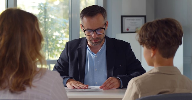 Vista posteriore della madre e del figlio durante il colloquio con il preside della scuola. (Adobe Stock)