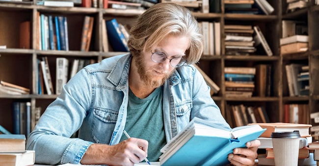 Giovane seduto al banco della biblioteca mentre prende appunti. (Alamy Stock Photo)