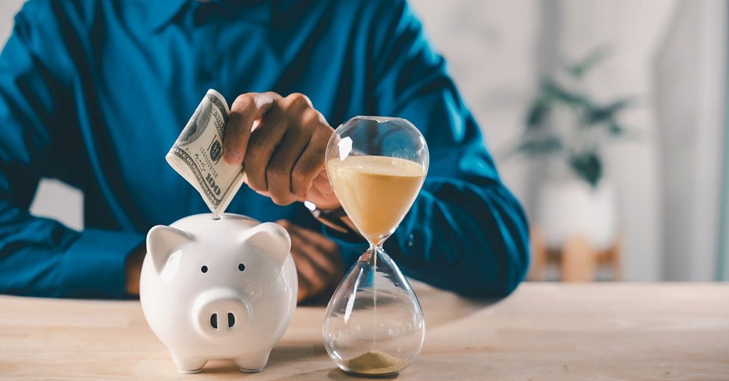 Hourglass money save cost company saving bank piggy business budget account time shows businessman putting hundred dollar bill into white ceramic piggy bank on wooden office desk.
