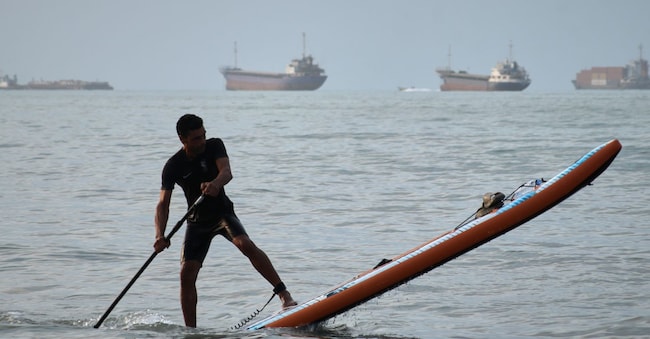 Un uomo su una tavola e sullo sfondo navi cargo ferme nello stretto di Hormuz  APN
