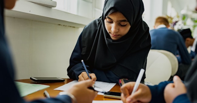 Studentesse musulmane con l'hijab nero intente a scrivere al banco, concentrate su un compito. Rawpixel.com - stock.adobe.com
