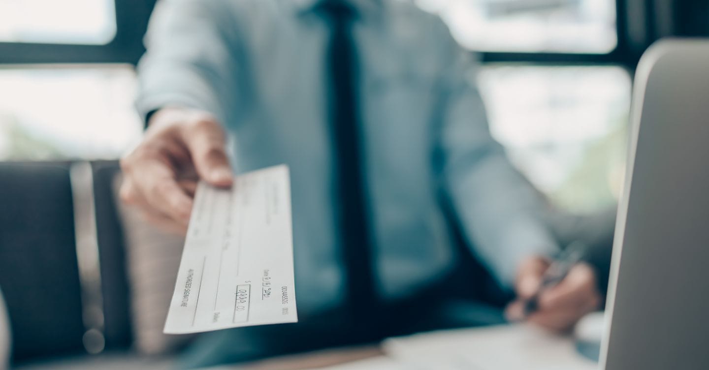 A Businessperson's hand giving cheque to customer and dollar bill, coin, laptop and graph chart on the desk at office. Payment by check, paycheck, payroll concept. Charnchai saeheng - stock.adobe.com